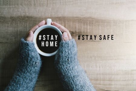 Woman's hands in gray sweater holding cup of black coffee with text hashtag #STAY HOME and #STAY SAFE on the wooden table. Concept stay home stop virus.の写真素材