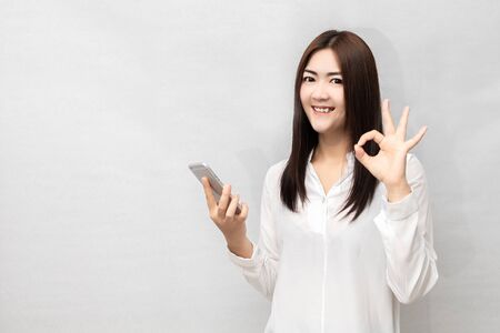 A long haired asian woman with a mobile phone while standing and showing ok gesture. News people concept. on white background.の写真素材
