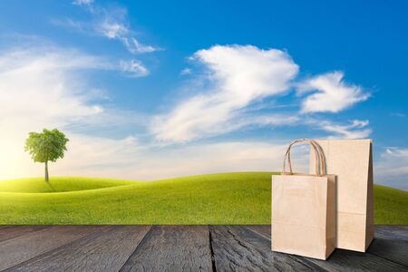 Recycle paper bag on old wooden floor beside green field on slope and tree with blue sky and clouds background.の写真素材