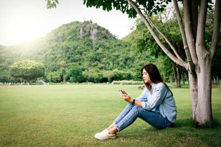 Woman using a smart phone resting on the grass in a park with warm tone.の写真素材