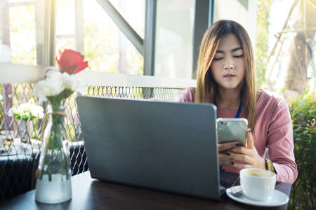 Asian woman using smart phone and laptop with white coffee cup in cafe.の写真素材
