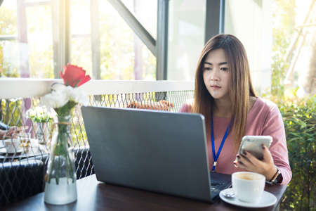 Asian woman using laptop computer and smart phone with white coffee cup on table in cafe.の写真素材