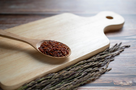 Red rice in wooden spoon on wooden cutting board and ears of rice on table.の写真素材
