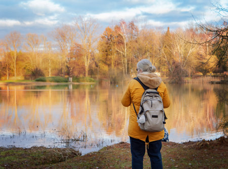A woman with a backpack in a yellow jacket looks at the lake on a sunny autumn day. Active recreation of the elderly. Nature and recreation. Braunschweig, Doweseeの写真素材