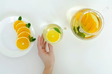 Female hands holding a cup of detox tea with orange fruit and mint on whitw background. Spa and...