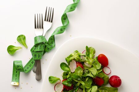 Fresh vegetable salad of radish, tomatoes and corn salad or Valerianella locusta. Close up on white plate and measuring tape wrapping forks. Diet, weight loss and slimming concept. Copy spaceの写真素材