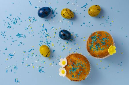 Muffins decorated with sprinkles, flowers of mastic and yellow and blue easter quail eggs on pastel blue background. Top view. Easter Holidays and greeting card concept.の写真素材