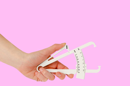 Female hand holding caliper on pink background. Copy space. Weight loss and slimming treatment...