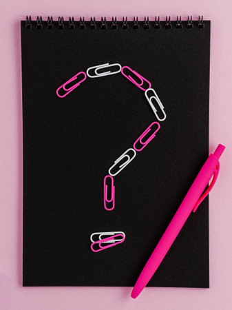 Question mark made of clips on black notebook blank on pink background. Flat lay.