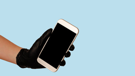 Hand in black glove holding smartphone with empty blank screen on blue background. Safe food...