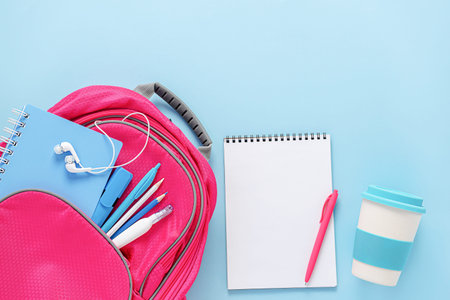 Pink backpack filled with stationery and reusable eco cup on blue background. Overhead, mockup...