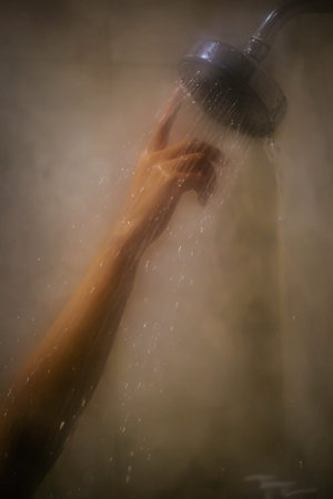 A hand reaches up under the stream of water from a showerhead, with droplets captured mid-fall in...