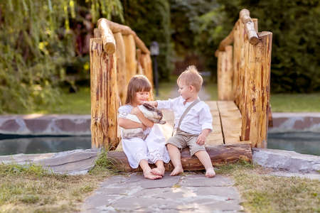 A little girl and a boy sit on a bridge over a river. A girl holds a pet rabbit in her hands. The boy smiles shyly. Nature walk.の写真素材