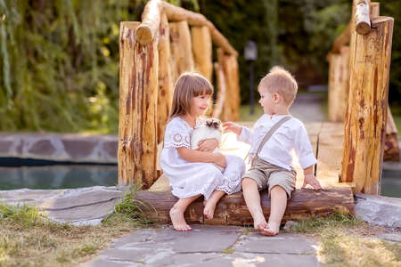 A little girl and a boy sit on a bridge over a river. A girl holds a pet rabbit in her hands. The boy wants to pet the rabbit. Nature walk.の写真素材
