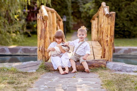 A little girl and a boy sit on a bridge over a river. A girl holds a pet rabbit in her hands. The boy wants to pet the rabbit. Nature walk.の写真素材