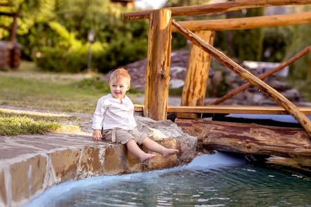 A little boy is sitting on the Bank of the river. Looks at the water. Swinging his legs in the water. Enjoys a walk in nature.の写真素材