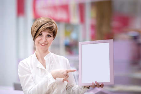 A beautiful young woman stands in a shopping center (or restaurant), holding a frame for ads. Copy space. Space for text. The concept of small business.の写真素材