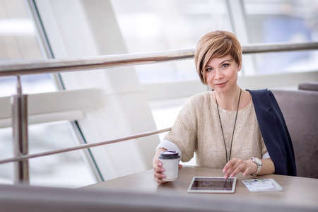 A young beautiful woman is sitting in a cafe, drinking coffee and browsing social media on a tablet. Lifestyle. Portrait of a woman watching social networks.の写真素材