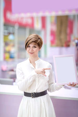 A beautiful young woman stands in a shopping center (or restaurant), holding a frame for ads. Copy space. Space for text. The concept of small business.の写真素材