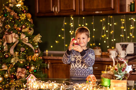 A boy sits at a table in a decorated Christmas kitchen. Holding a toy for decorating a Christmas tree. Cozy winter evenings at home. Concept of Christmas and new year holidays at home. selective focus; contour light; shallow depth of fieldの写真素材