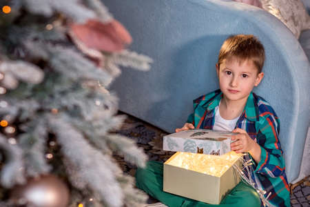 A boy is sitting on the floor near a decorated Christmas tree. Opens a box with a glowing garland. Cozy winter evenings at home. Concept of Christmas and new year holidays at home. selective focus; contour light; shallow depth of fieldの写真素材
