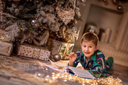 The boy is lying on the floor near a decorated Christmas tree. Reading a book with Christmas stories. Cozy winter evenings at home. Concept of Christmas and new year holidays at home. selective focus; contour light; shallow depth of fieldの写真素材