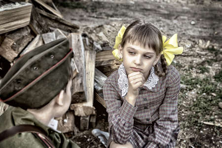 Postcard, stylized as vintage for the Victory Day. A boy in a military uniform and a girl in an old dress are sitting on stumps near a woodpile. The theme of May 9, Victory Day in Russia. Soft selective focus, added noiseの写真素材