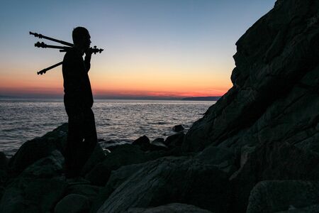 Dark silhouette of a man holding a tripod on his shoulder against the backdrop of a sea sunsetの写真素材