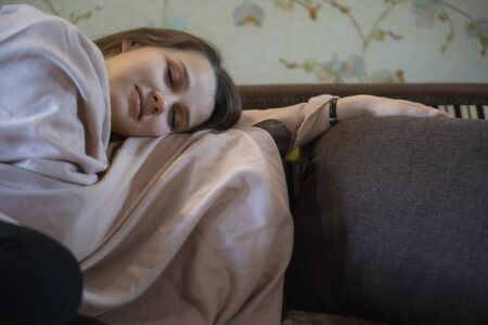A picture of nice and thoughtful girl sits at sofa and reads a book. She has brown blanket on her shoulders. Woman got cold. She suts at windowの写真素材