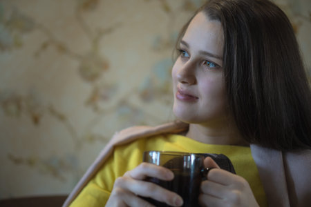 A picture of nice and thoughtful girl sits at sofa and drinks tea. She has brown blanket on her...