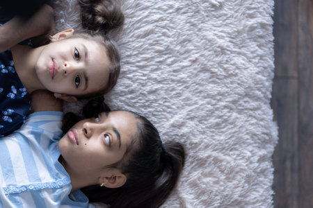 Two Middle Eastern young cute girls, sisters, lie on the bed and look at the smartphone in the rays...