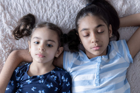 Two Middle Eastern young cute girls, sisters, lie on the bed and look at the smartphone in the rays...