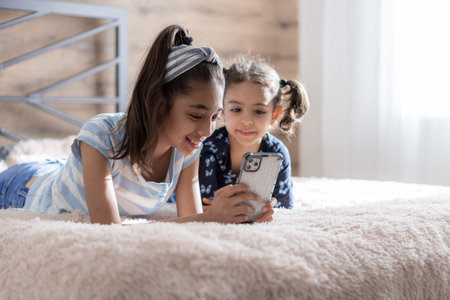 Two dark-skinned young girls, sisters, are lying in bed and looking at a smartphone against the background of the sun from the window. Persian girls on the bed with a phone. Middle Eastern kids.の写真素材