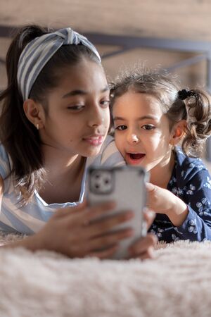 Two dark-skinned young girls, sisters, are lying in bed and looking at a smartphone against the background of the sun from the window. Persian girls on the bed with a phone. Middle Eastern kids.の写真素材