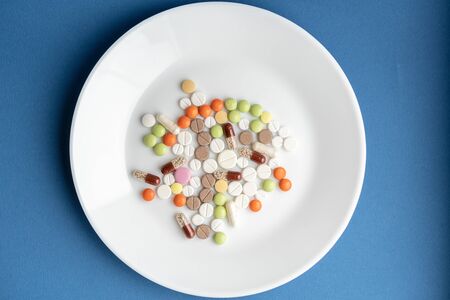 Pills on a white plate. Multi-colored pile of pills on a classic blue background. Medication on a white table. Pills under the guise of food on a plate. A bunch of pills symbolizing food.の写真素材