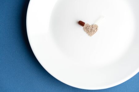 Granules poured from a capsule onto a white plate in the shape of a heart on a classic blue background. The heart symbol is made of granules spilling out of a capsule on a blue background.の写真素材
