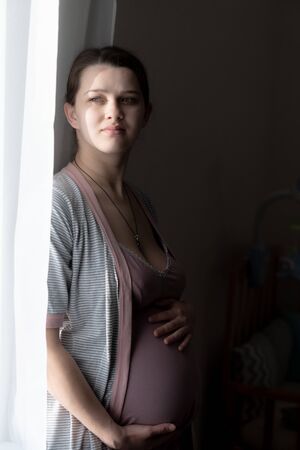 A cute pregnant young woman is looking through a curtain through a sunny window. A pregnant girl in a nightie is standing near the window and smiling.の写真素材