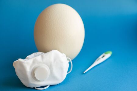 Large white easter ostrich egg in a respirator and a thermometer. Egg, respirator and thermometer. Easter and Coronavirus, Covid-19. Ostrich egg, medical mask and thermometer on a classic blue background.の写真素材