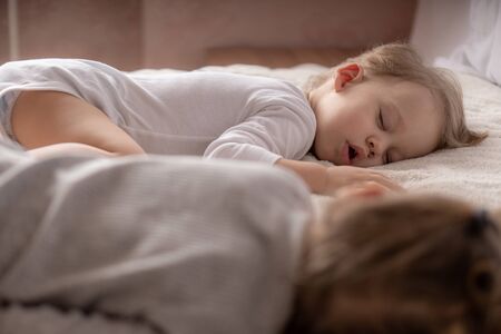 Childhood, sleep, relaxation, family, lifestyle concept - two young children 2 and 3 years old dressed in white and beige bodysuit sleep on a beige and white bed at lunch holding hands top viewの写真素材