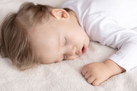 Childhood, sleep, rest, family, lifestyle concept - close-up portrait of a cute little boy of 2 years old in a white body sleeping on a beige bed at noon with mouth open top and side view.の写真素材
