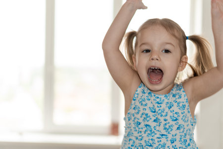 childhood, people, emotions concept - medium plan little girl with two ponytails and a blue dress jumps raises her hands and screams at the window.の写真素材