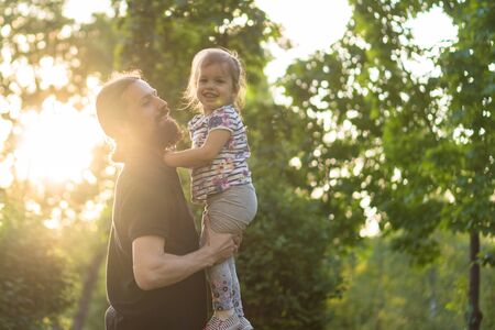 Fatherhood, parenthood, childhood, caring, summer and leisure concept - young dad with beard and long hair in black t-shirt holding his little daughter in his arms in backlight of sunset in the park.の写真素材