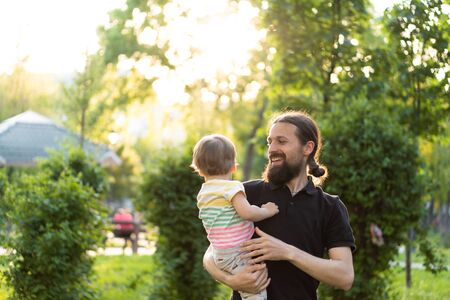 Fatherhood, parenthood, childhood, caring, summer and leisure concept - young dad with beard and long hair in black t-shirt holds in his arms little son in the backlight of the sunset in the park.の写真素材