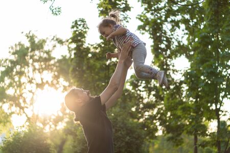 Fatherhood, parenthood, childhood, caring, summer and leisure concept - young dad with beard and long hair in black t-shirt throws up her little daughter in the backlight of the sunset in the park.の写真素材