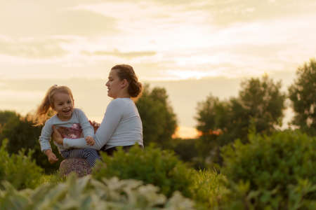 Motherhood, care, infants, summer, parenting concept - Young mom with with three year old little girl daughter in her arms walks and looks at child in backlight of sunset in park near flowering bush.の写真素材