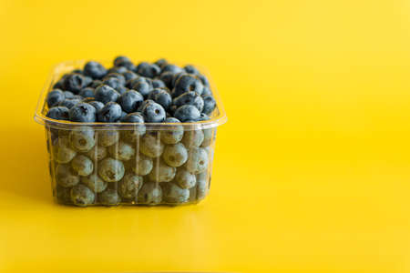 fruit and oat, vitamin, healthy food and drink, traditional medicine, gastronomy concept - purple and blue blueberry berries in plastic charcoal transparent tray on bright yellow background copy spaceの写真素材