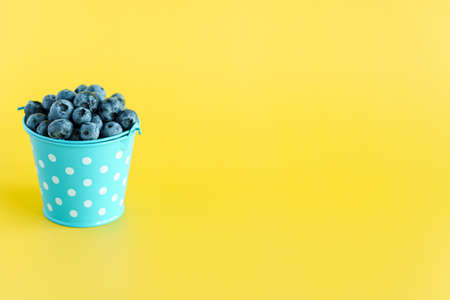 fruits and vegetables, vitamin, healthy food and drink, traditional medicine, gastronomy concept - layout purple blueberry berries in blue bucket with white dots on bright yellow background copy spaceの写真素材