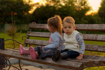 infancy, childhood, development, autumn mood, medicine and health concept - two small children boy and girl of same age thoughtfully sitting on park bench waiting on green meadow in park at sunset.の写真素材