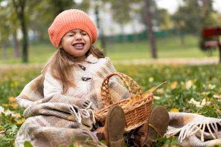Childhood, family, infant, motherhood, autumn concept - Little cute preschool minor blonde girl in orange beret sits on plaid with basket and yellow fallen maple leaves laughs smiling with closed eyesの写真素材