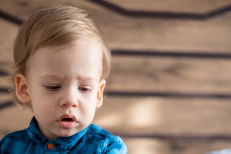 childhood, infant, minor concepts - close up one blonde black-eyed sad, offended child baby boy on soft powdery bed in sunny room. portrait embarrassed preschool Slavic kid in shirt at home indoorsの写真素材